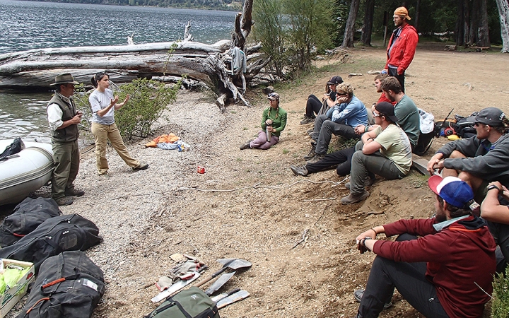 A group of people sit on the shore, listening to two people speak. In the water, there is a raft.